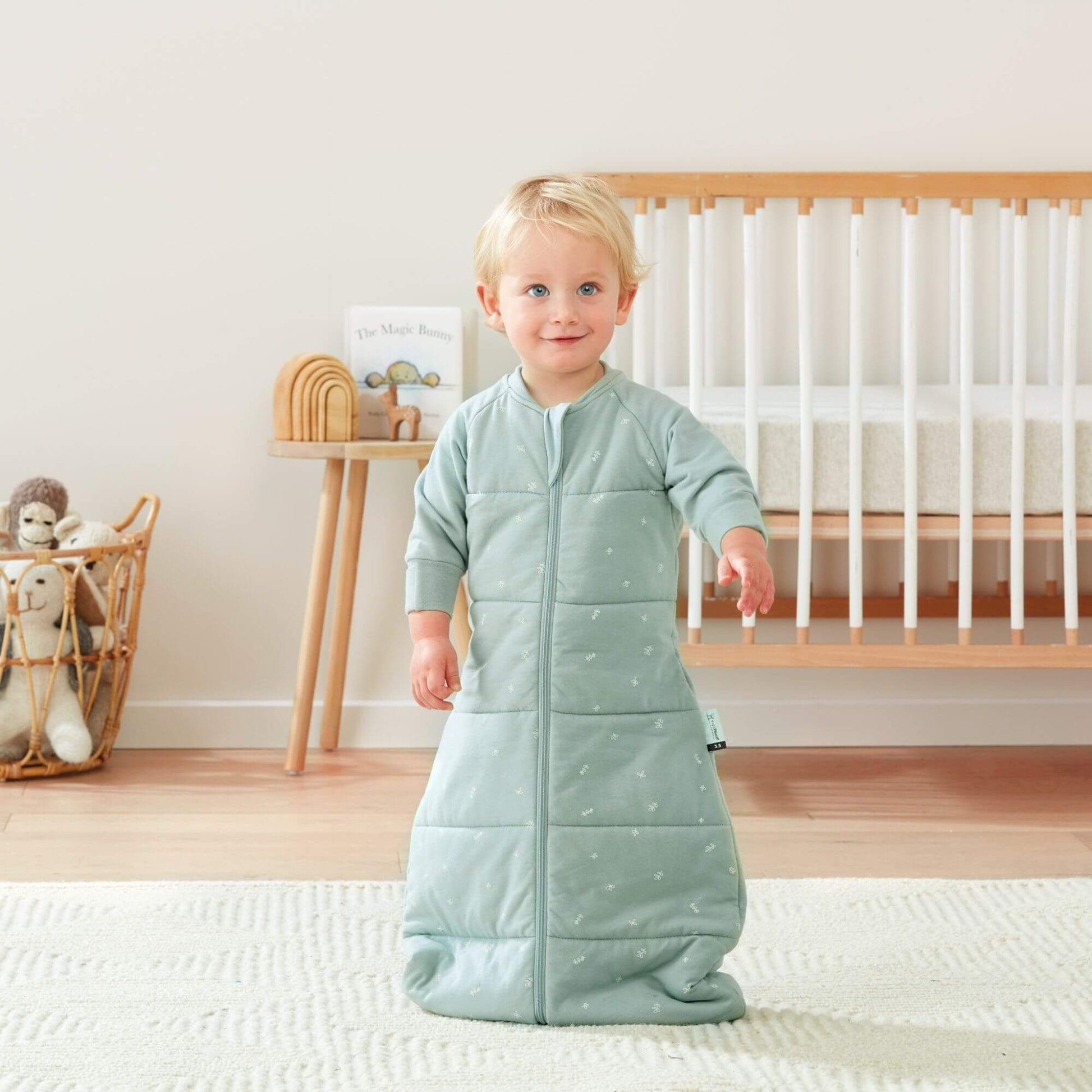  A toddler stands wearing a sage green sleeping bag, in a nursery setting with a cot and toys nearby.