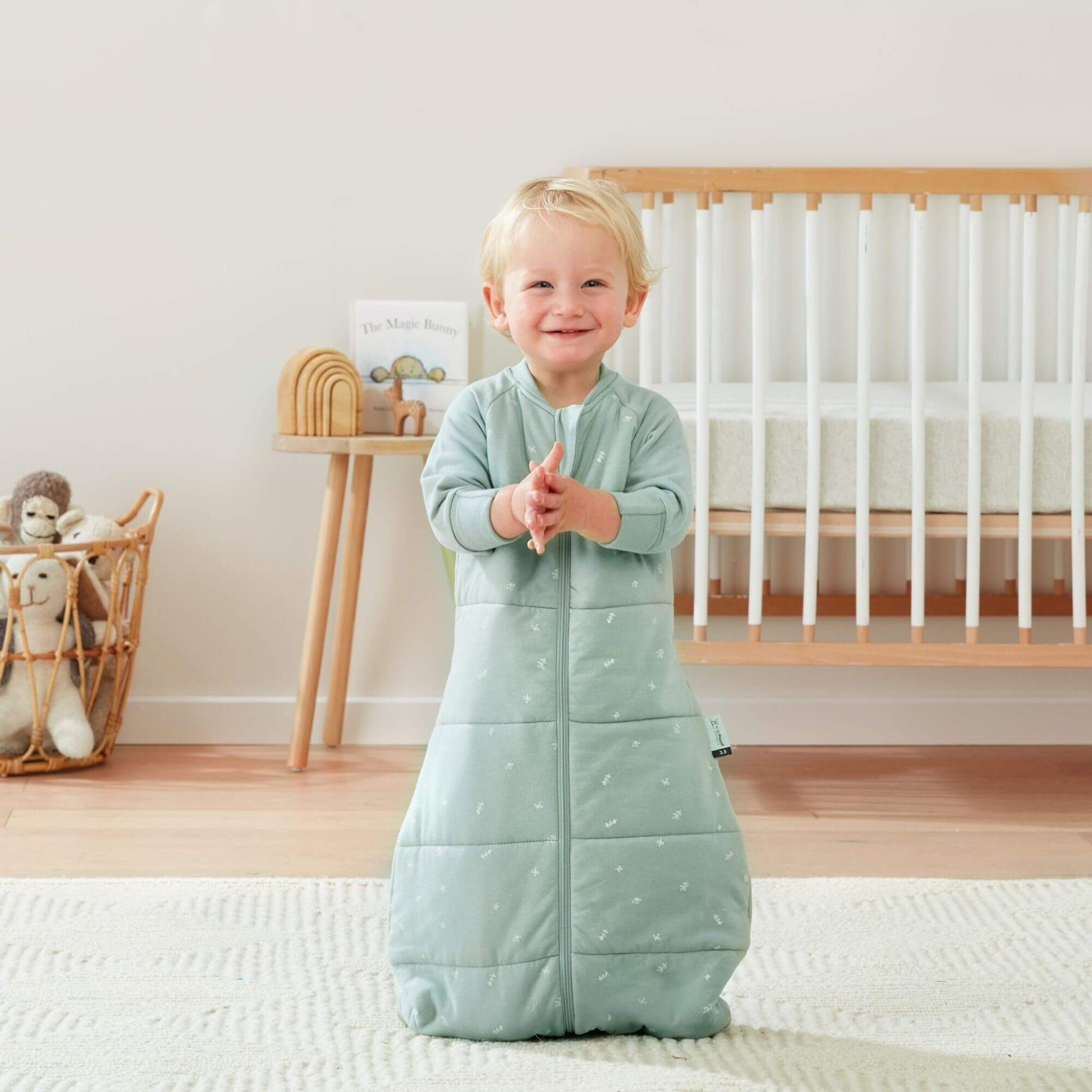 A smiling toddler claps while wearing a sage green sleeping bag, in a nursery with a cot and toys.