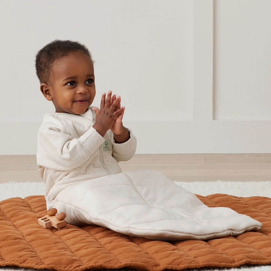 Baby sitting happily in a Long Sleeved Sleeping Bag in Oatmeal, with a cosy zip-up design and bell-shaped bottom.