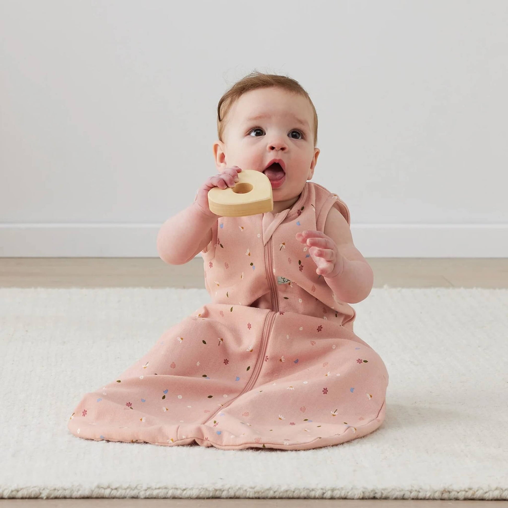 Baby sitting upright on a cream rug wearing a pink sleeveless sleeping bag decorated with small daisies, holding a wooden heart-shaped toy and looking upward.