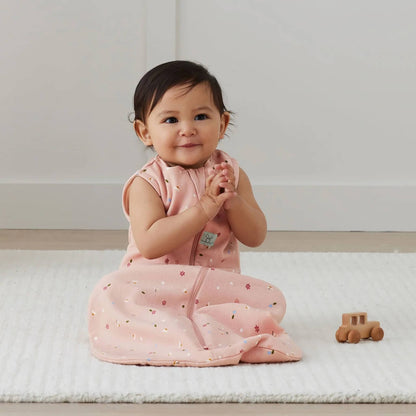 Baby sitting on a cream rug in a pink sleeveless sleeping bag with a daisy pattern, smiling with hands together while a small wooden toy car rests beside them.