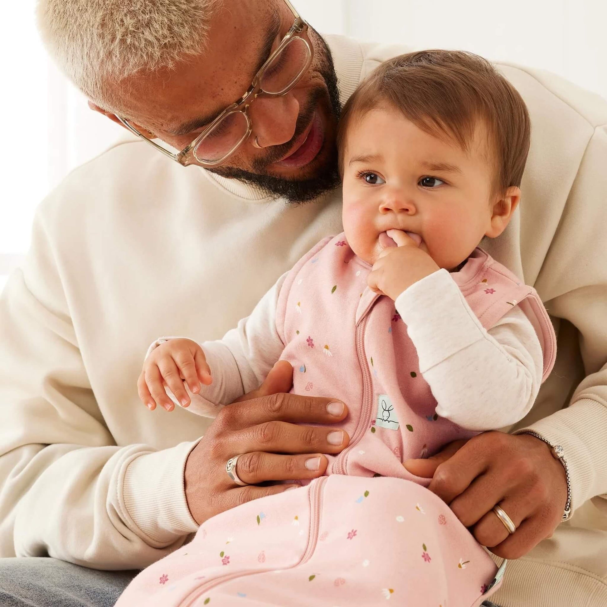 Adult holding a baby on their lap; the baby wears a pink floral sleeping bag and long-sleeved top, with fingers in their mouth while leaning against the adult’s chest.