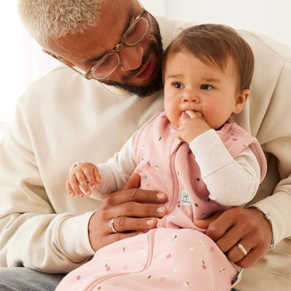 Adult holding a baby on their lap; the baby wears a pink floral sleeping bag and long-sleeved top, with fingers in their mouth while leaning against the adult’s chest.