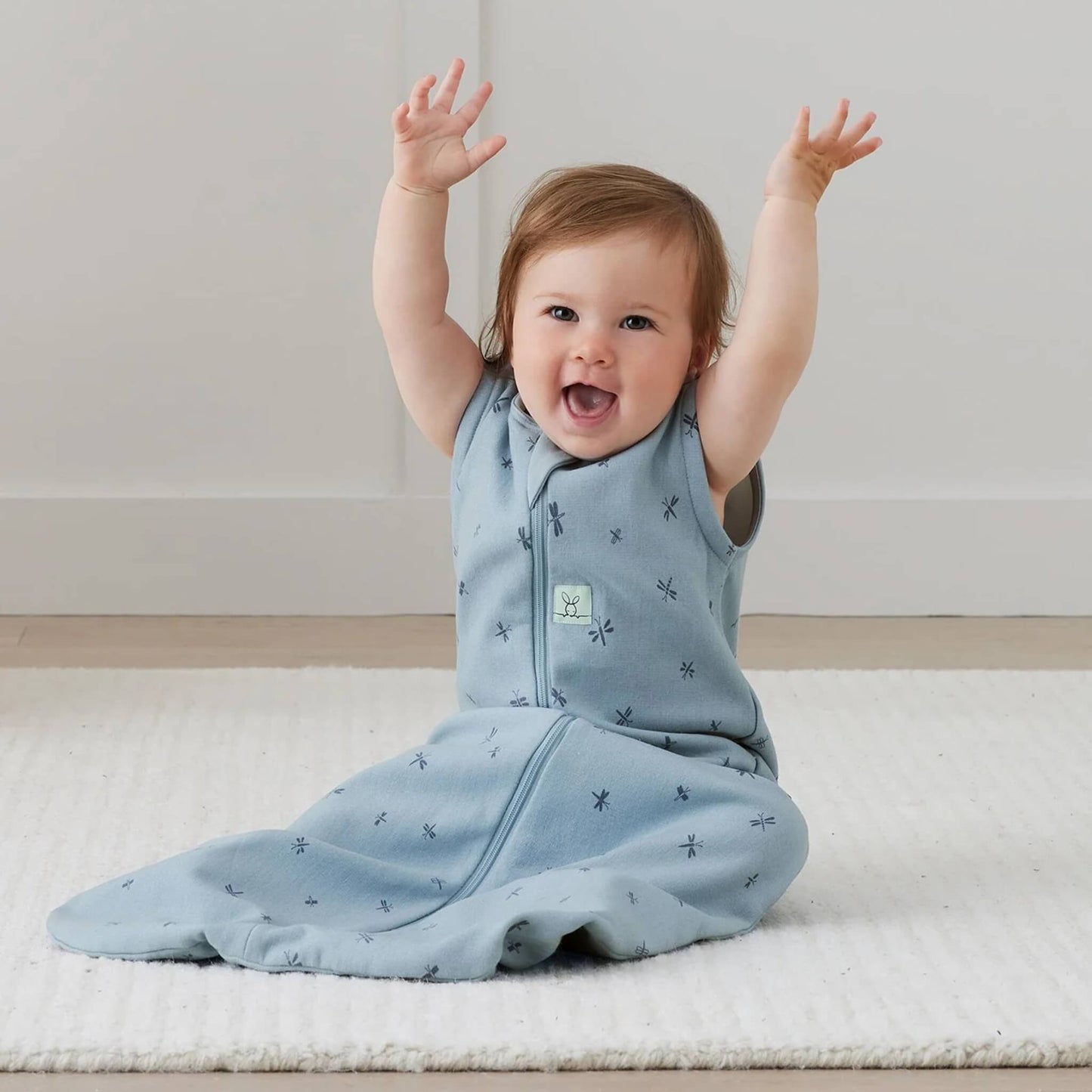 Smiling baby sitting on a light rug in a sleeveless blue sleeping bag with dragonfly pattern, arms raised playfully in the air.
