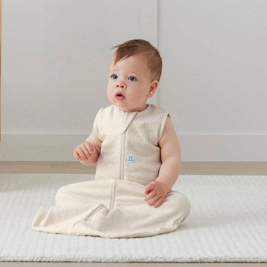 Baby sitting upright on a cream rug wearing a sleeveless oatmeal-coloured sleeping bag, looking upward with hands resting on their lap.