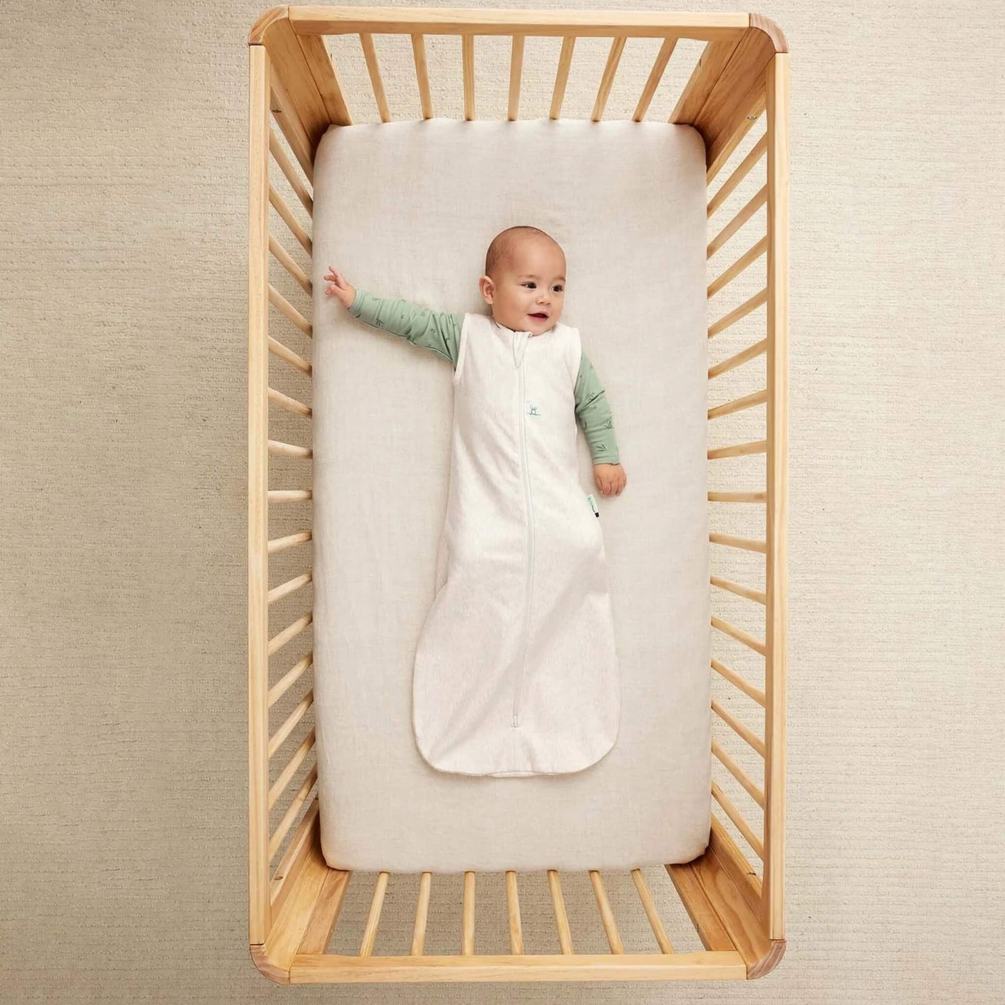 Baby lying on their back in a wooden cot, dressed in a cream sleeveless sleeping bag over a green long-sleeved top, one arm stretched outward.