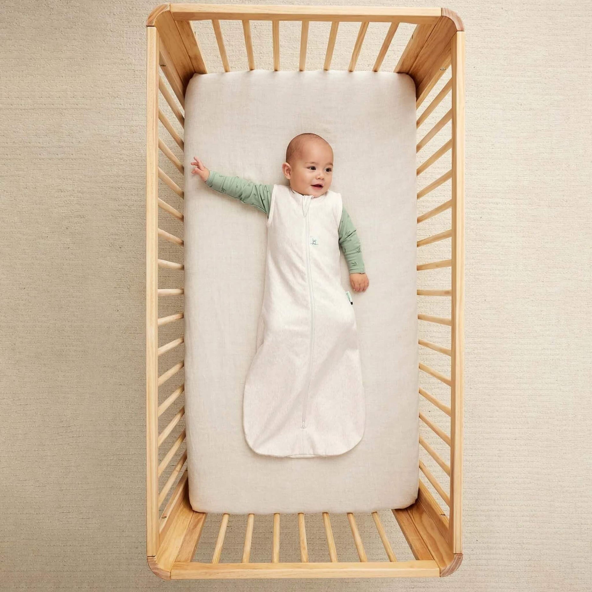 Baby lying on their back in a wooden cot, dressed in a cream sleeveless sleeping bag over a green long-sleeved top, one arm stretched outward.