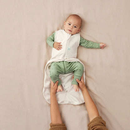 Baby lying on a beige surface wearing a cream sleeveless sleeping bag, with a green outfit underneath and a parent’s hands gently holding their legs for a nappy change.