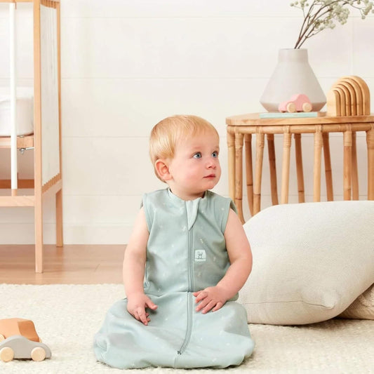 Toddler sitting upright on a light rug in a sleeveless pale green sleeping bag, looking to the side with a wooden toy car nearby.