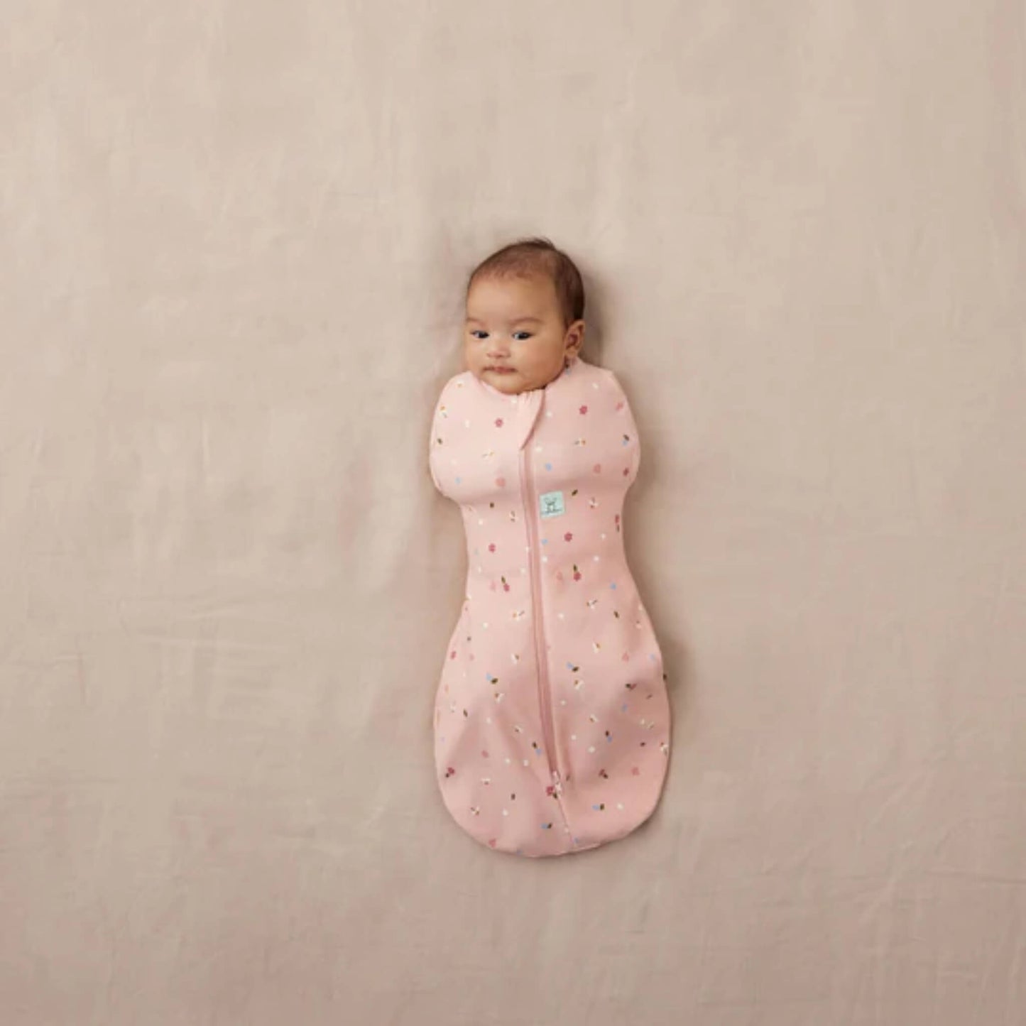 Newborn baby lying on a neutral surface wrapped in a pink Cocoon Swaddle Bag with daisy and ladybird print, arms fully swaddled inside.