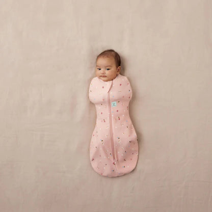 Newborn baby lying on a neutral surface wrapped in a pink Cocoon Swaddle Bag with daisy and ladybird print, arms fully swaddled inside.