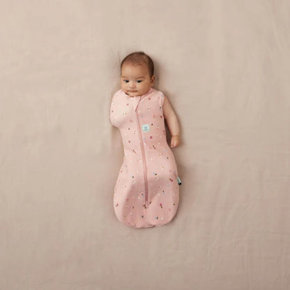 Baby lying on a neutral background wearing a pink Cocoon Swaddle Bag with daisy and ladybird print, one arm free and the other swaddled.