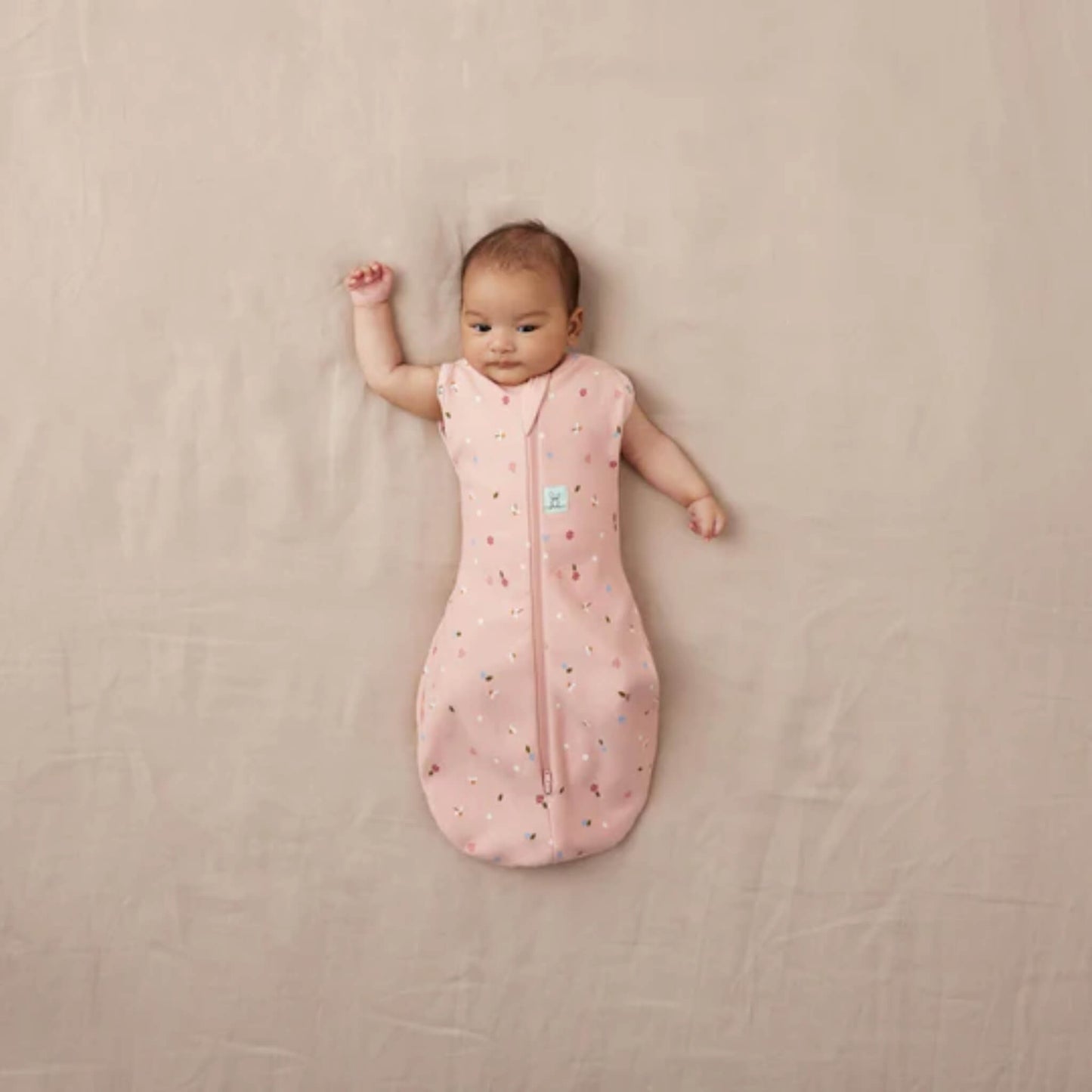 Baby resting on a neutral background in a pink Cocoon Swaddle Bag with daisy and ladybird design, both arms free.