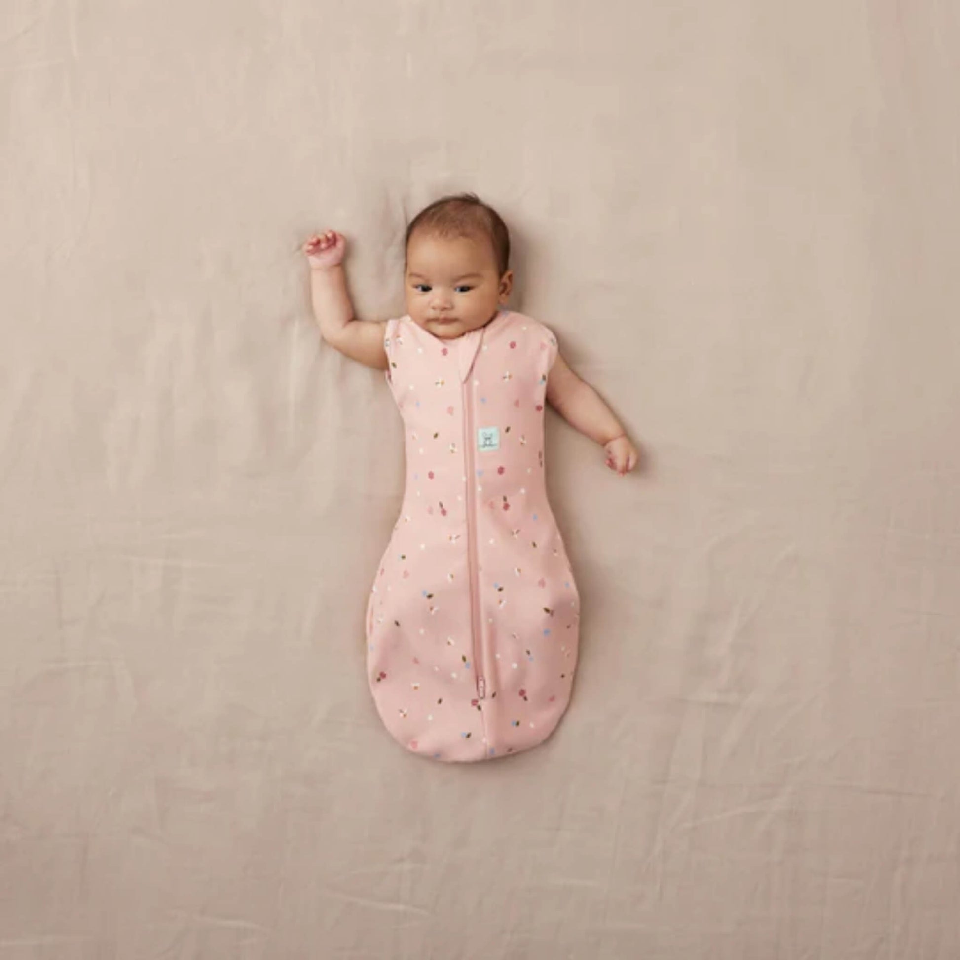 Baby resting on a neutral background in a pink Cocoon Swaddle Bag with daisy and ladybird design, both arms free.