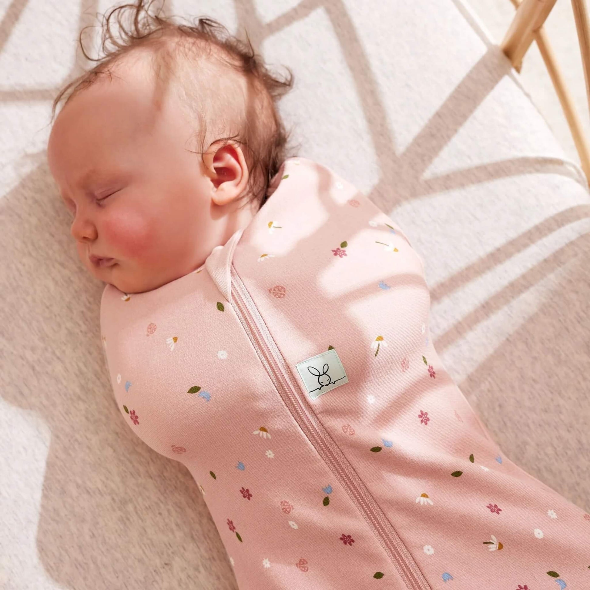 Close-up of a sleeping baby in a pink Cocoon Swaddle Bag with small daisies, flowers, and ladybird motifs.