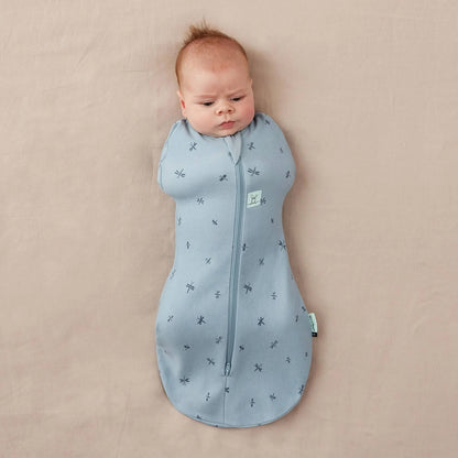 Baby lying on a neutral surface swaddled in a light blue Cocoon Swaddle Bag decorated with dragonfly print, both arms tucked inside.