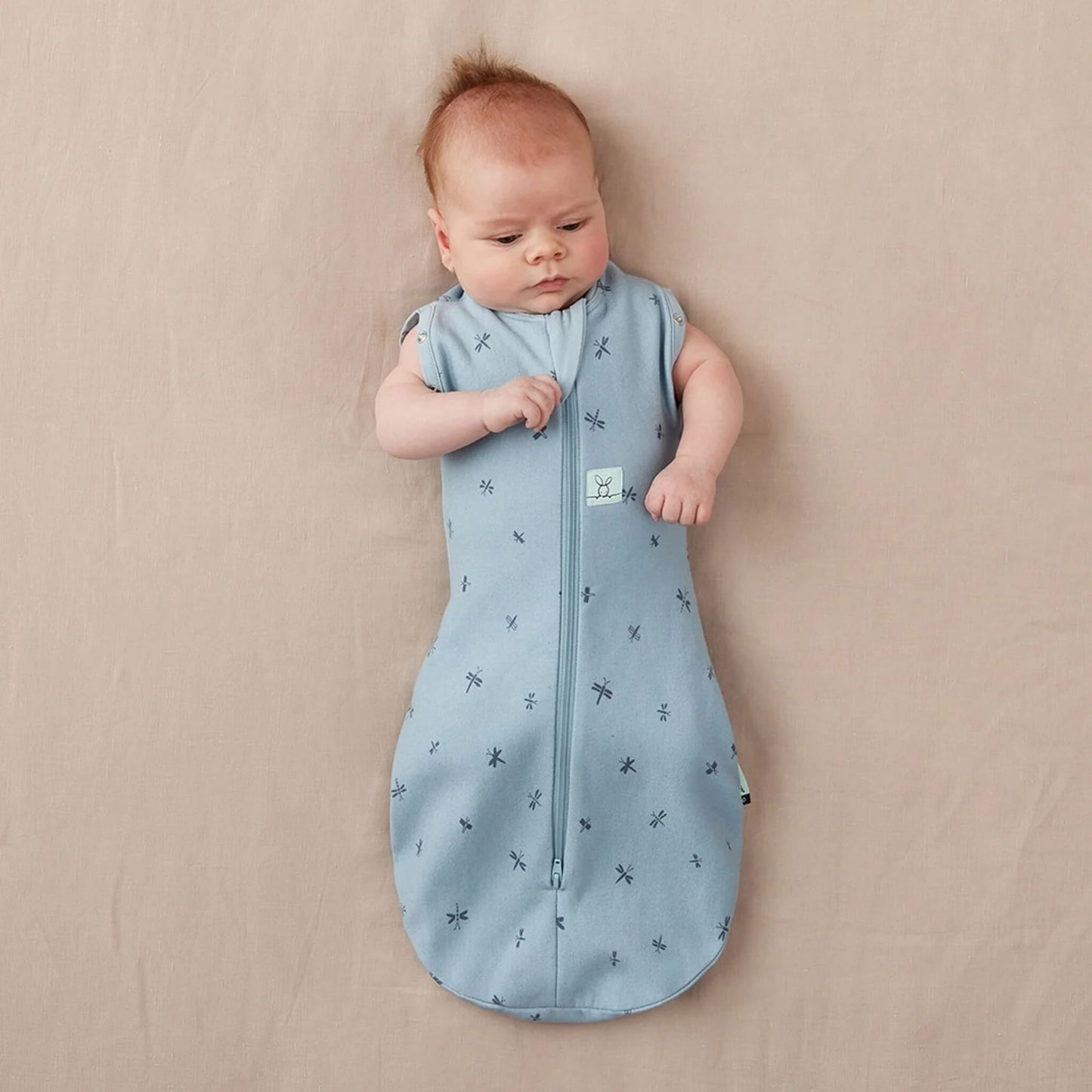 Baby lying on a neutral background wearing a light blue Cocoon Swaddle Bag with dragonfly design, both arms free.