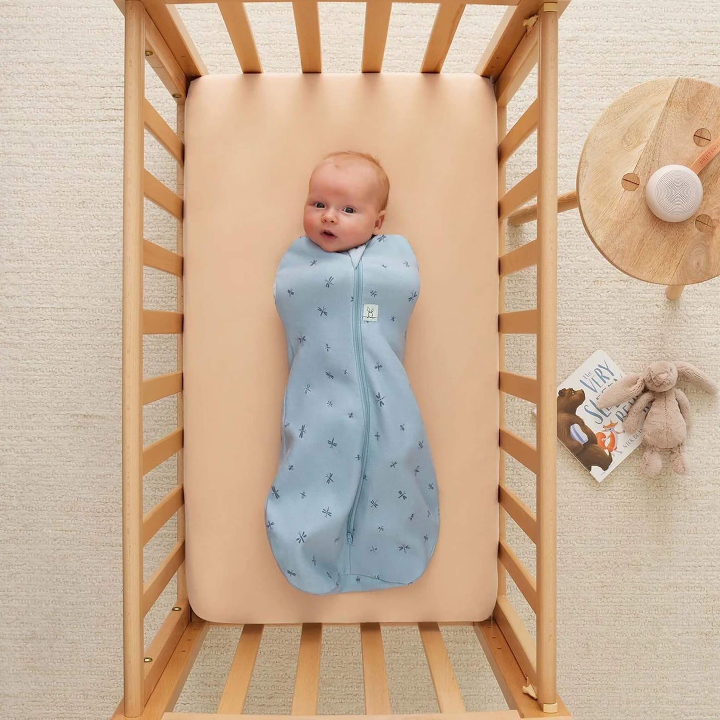 Baby lying in a wooden cot swaddled in a light blue Cocoon Swaddle Bag with dragonfly print, with a soft toy and book nearby.