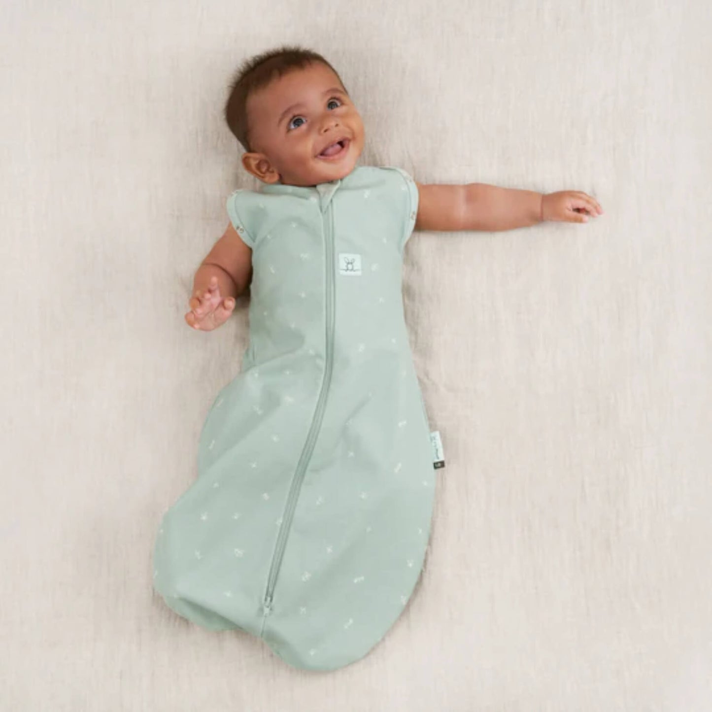 Baby resting with both arms out in a sage green Cocoon Swaddle Bag, worn as a sleeveless baby sleeping bag.