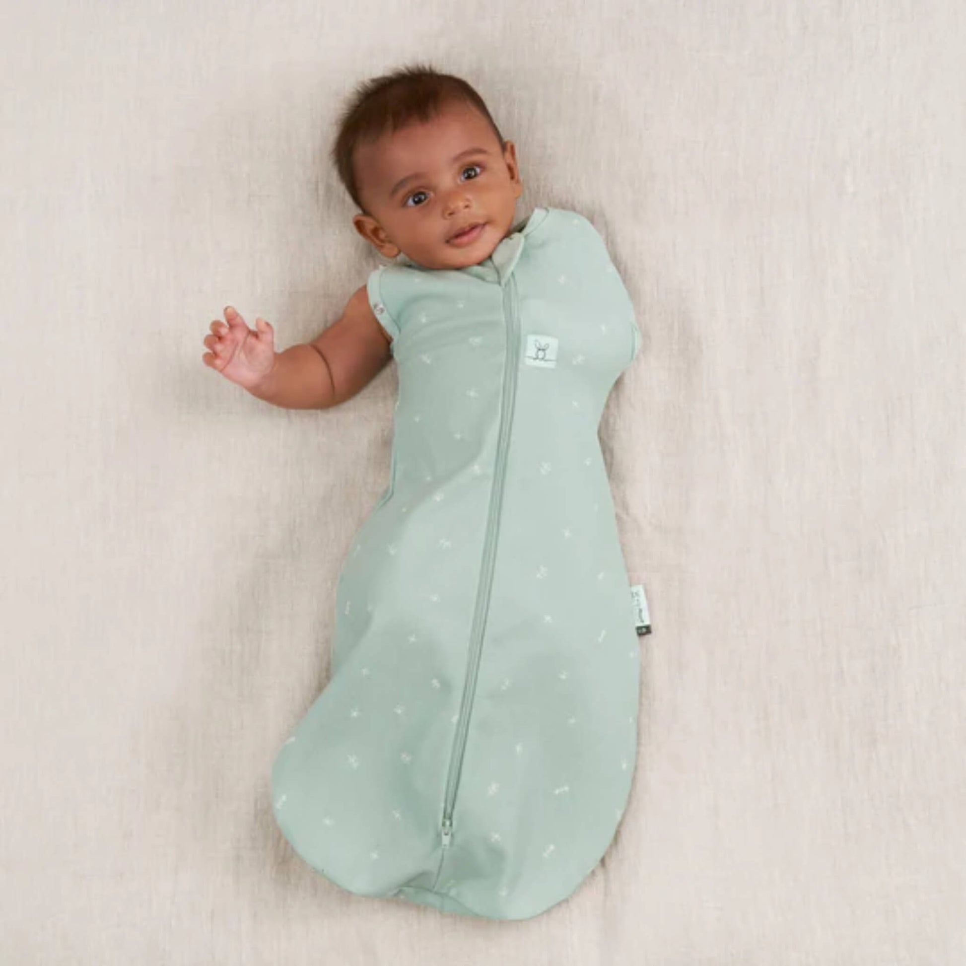 Smiling baby with one arm free, dressed in a sage green cotton swaddle bag that can transition into a sleeping bag.
