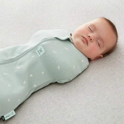 Sleeping baby shown close-up wearing a sage green swaddle bag with a central zip and white leaf design on soft cotton fabric.