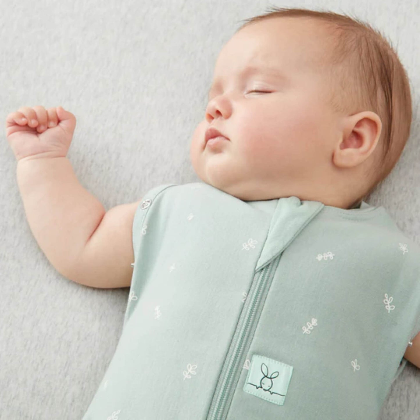 Baby sleeping with arm raised while wearing a sleeveless sage green Cocoon Swaddle Bag with front zip.