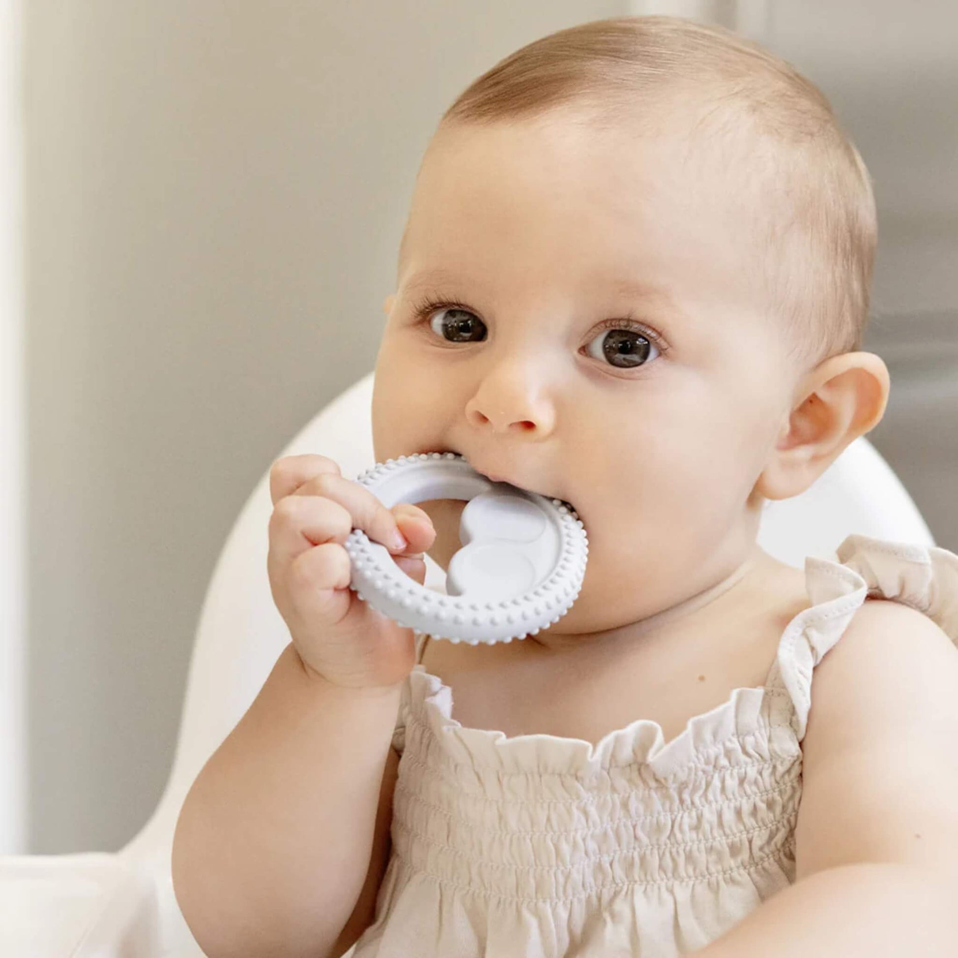 A baby holding and chewing a grey silicone oral development ring with raised sensory bumps designed for early oral motor exploration.