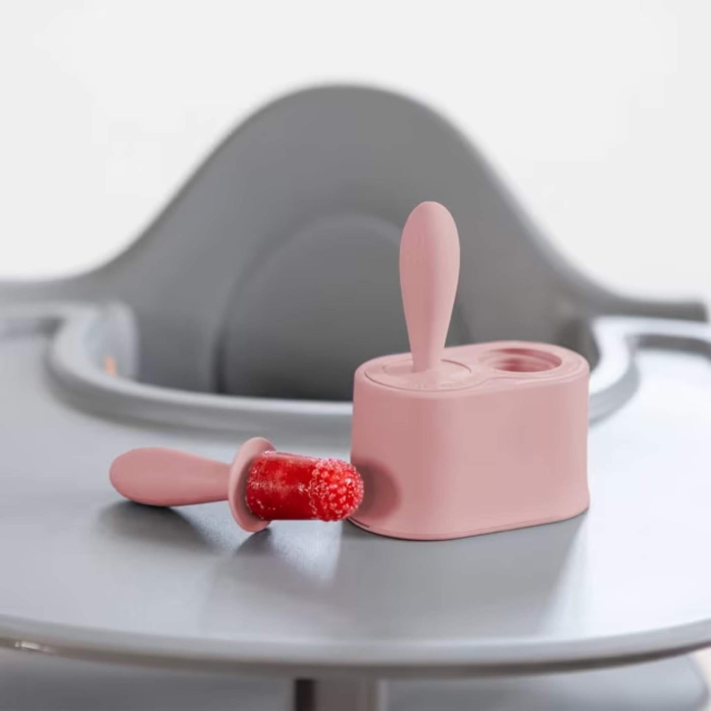 Blush silicone baby popsicle mould with frozen fruit pop beside stand placed on a highchair tray.