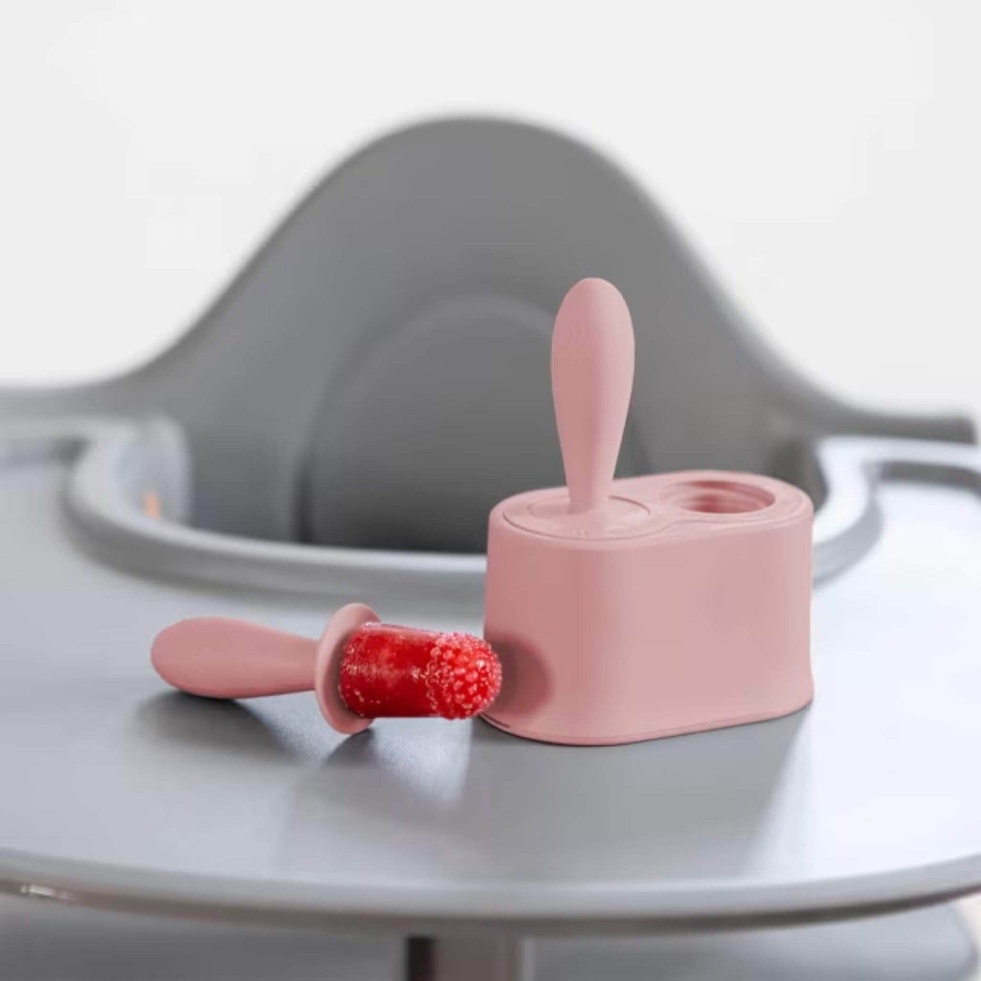 Blush silicone baby popsicle mould with frozen fruit pop beside stand placed on a highchair tray.