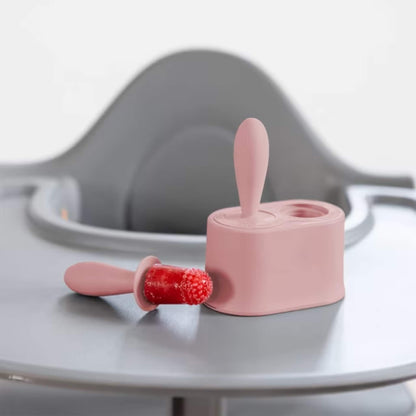Blush silicone baby popsicle mould with frozen fruit pop beside stand placed on a highchair tray.
