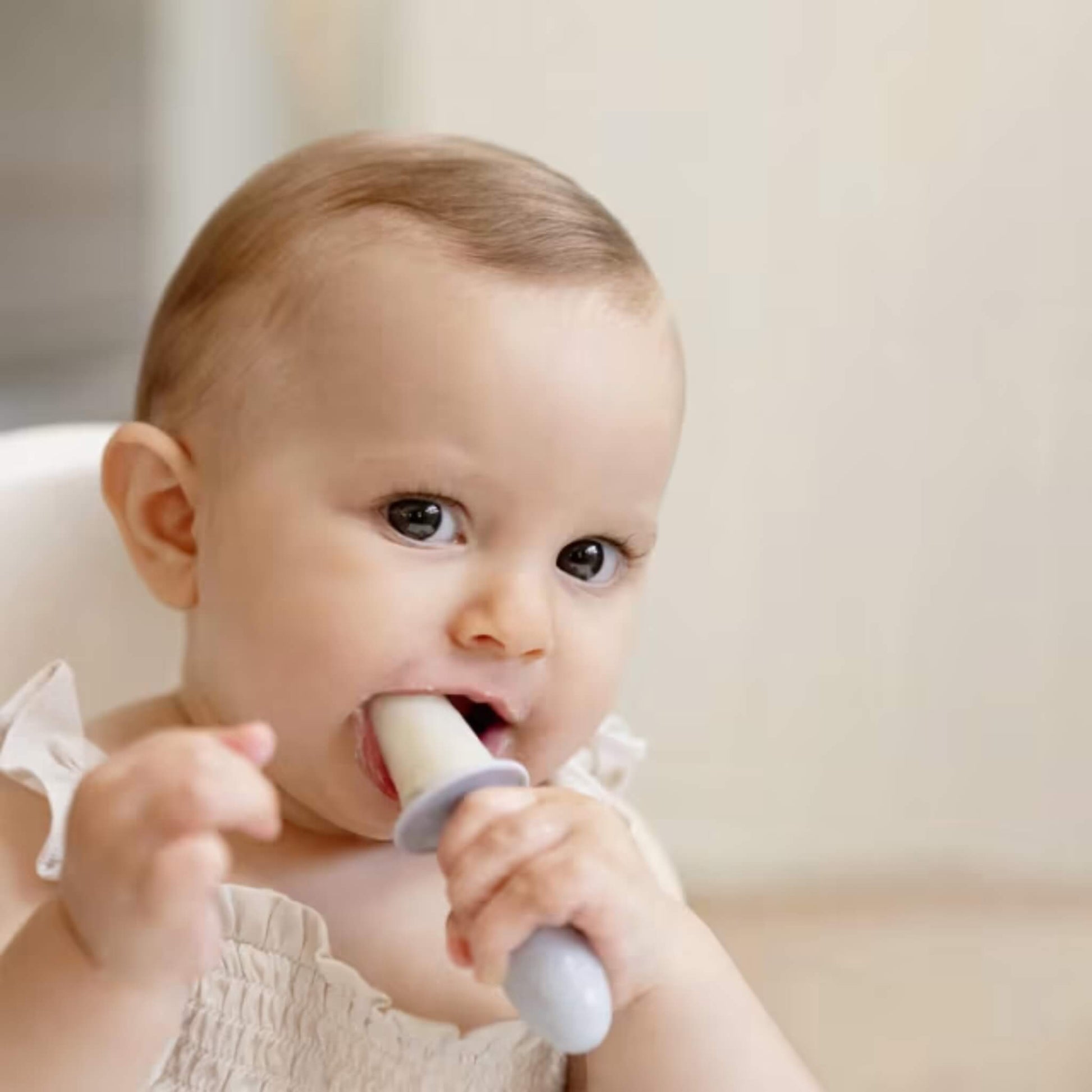 Baby sitting in a highchair enjoying a frozen purée popsicle designed for early self-feeding and teething comfort.