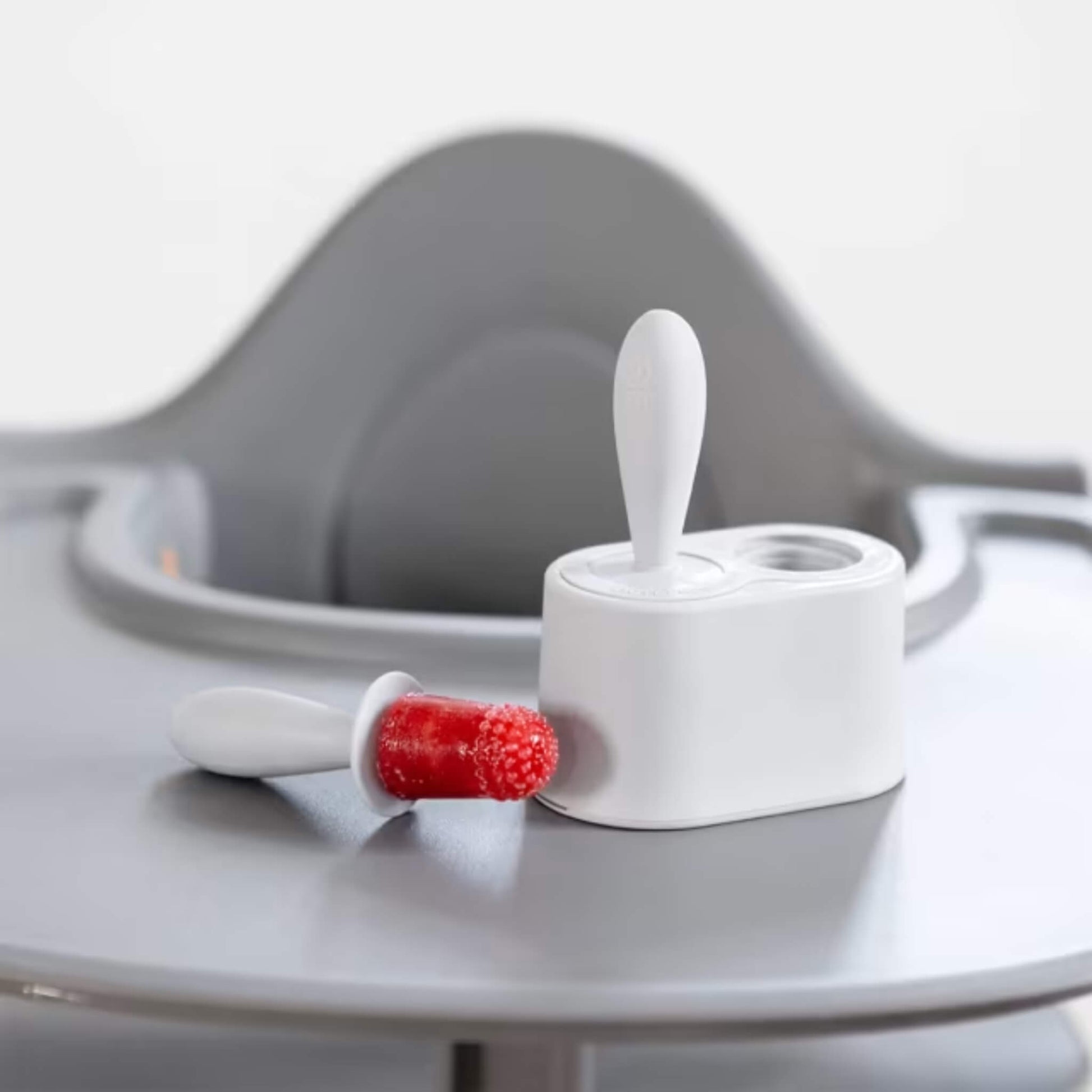 Frozen fruit baby popsicle with textured tip resting beside silicone mould stand on a highchair tray.