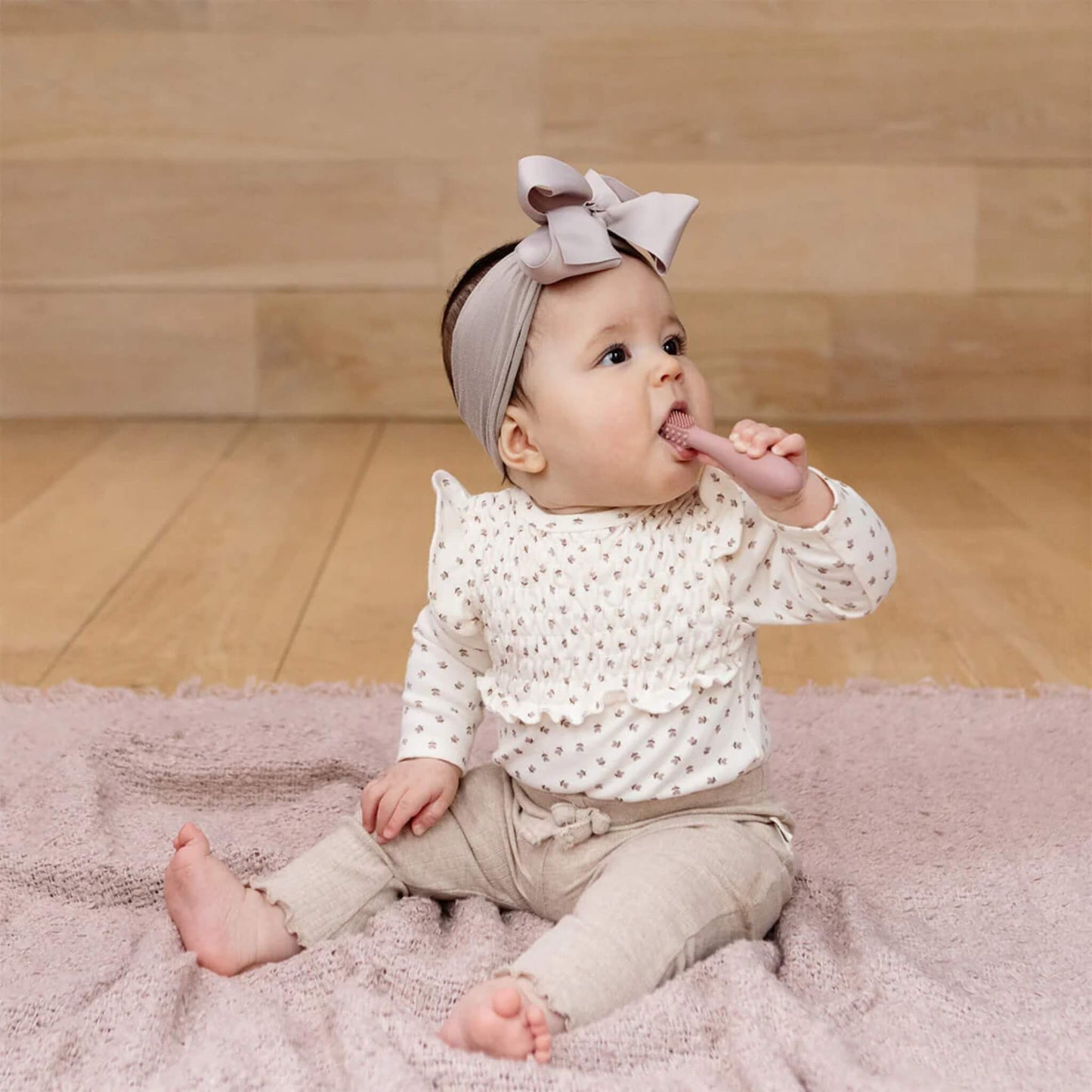 A baby sitting on a soft pink blanket holding a blush silicone toothbrush and chewing on the textured head while exploring early brushing.