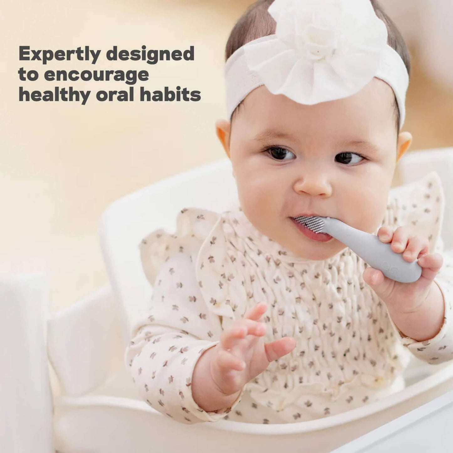 A baby sitting in a highchair holding a grey silicone toothbrush and chewing on a textured head while exploring early brushing and oral development.
