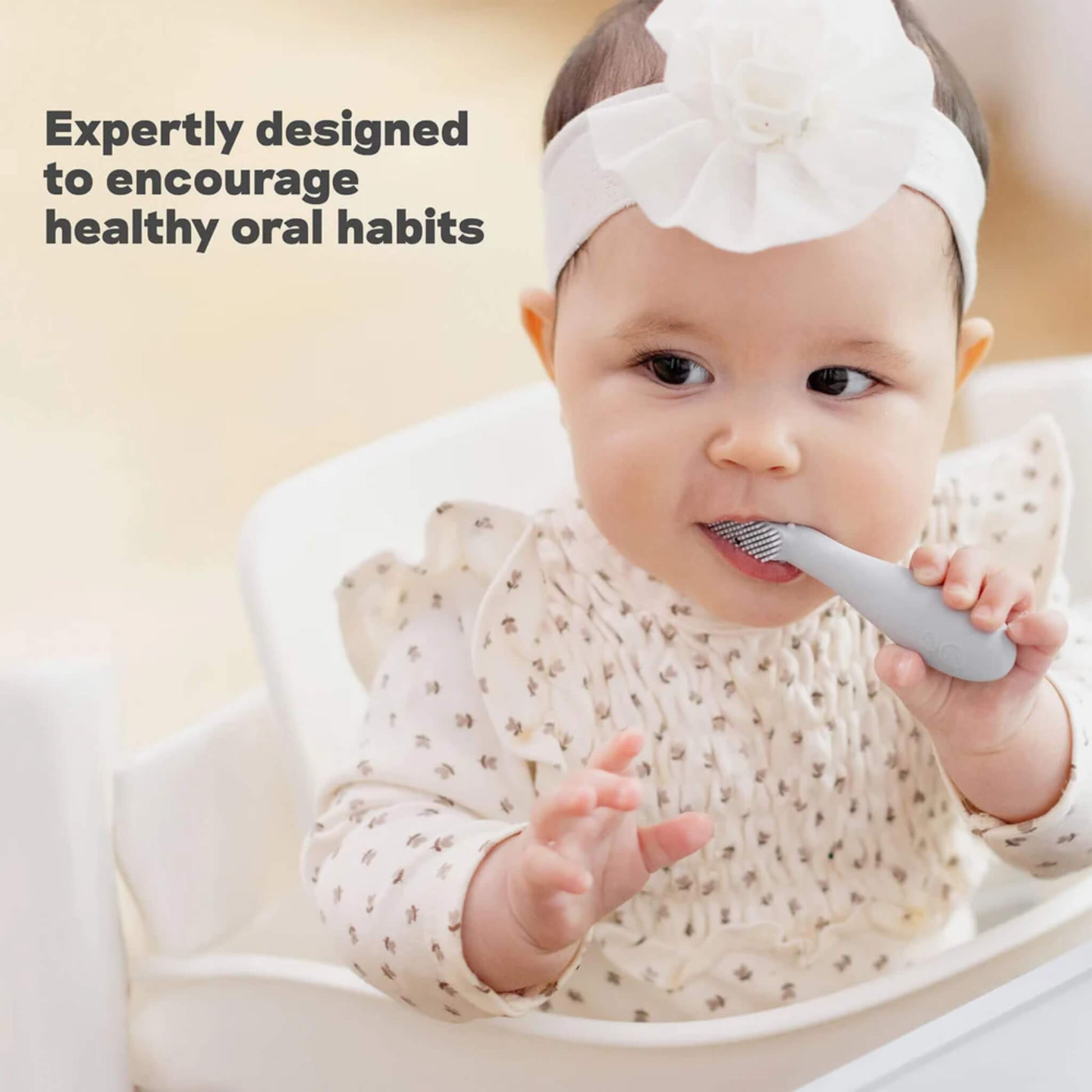 A baby sitting in a highchair holding a grey silicone toothbrush and chewing on a textured head while exploring early brushing and oral development.