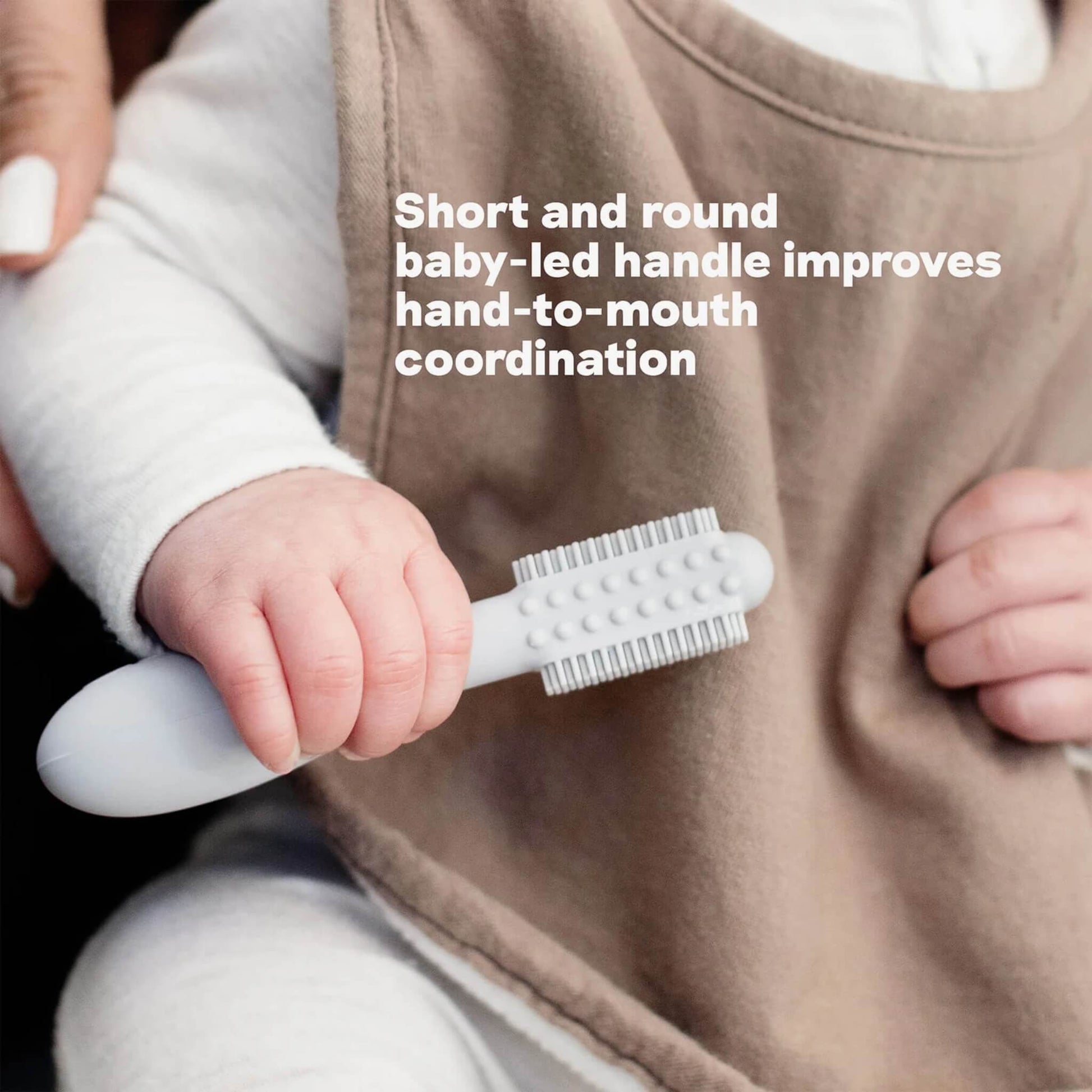 A baby hand holding a grey silicone toothbrush with short rounded handle and dual-sided bristles designed to support hand-to-mouth coordination.