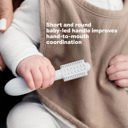 A baby hand holding a grey silicone toothbrush with short rounded handle and dual-sided bristles designed to support hand-to-mouth coordination.