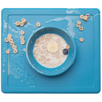 Blue silicone toddler placemat with a built-in bowl holding cereal, milk, banana slices and a blueberry. Milk and cereal pieces are spilled across the mat surface.