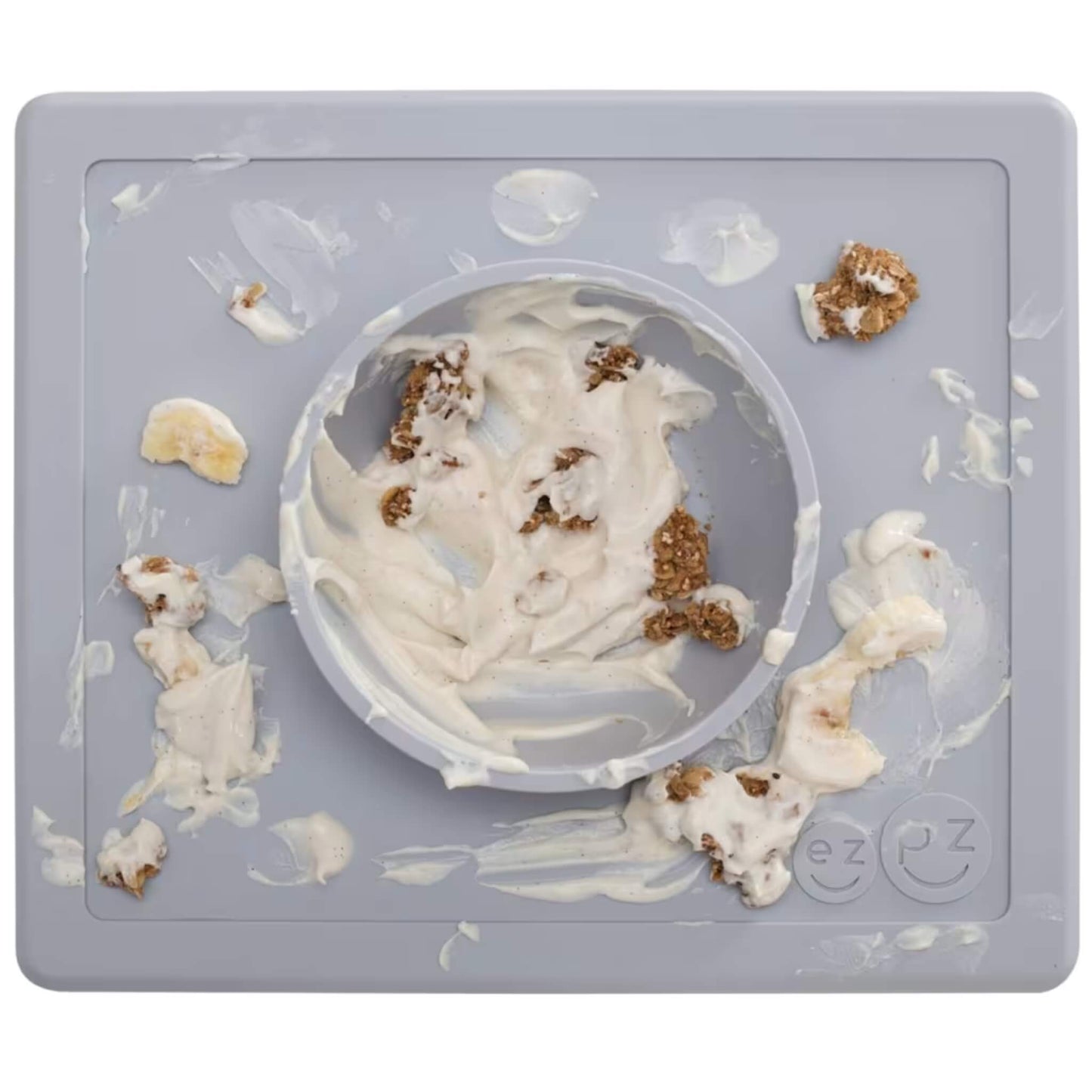 Grey silicone placemat with built-in bowl, shown messy with yoghurt, granola chunks and banana pieces spread across the mat and inside the bowl.