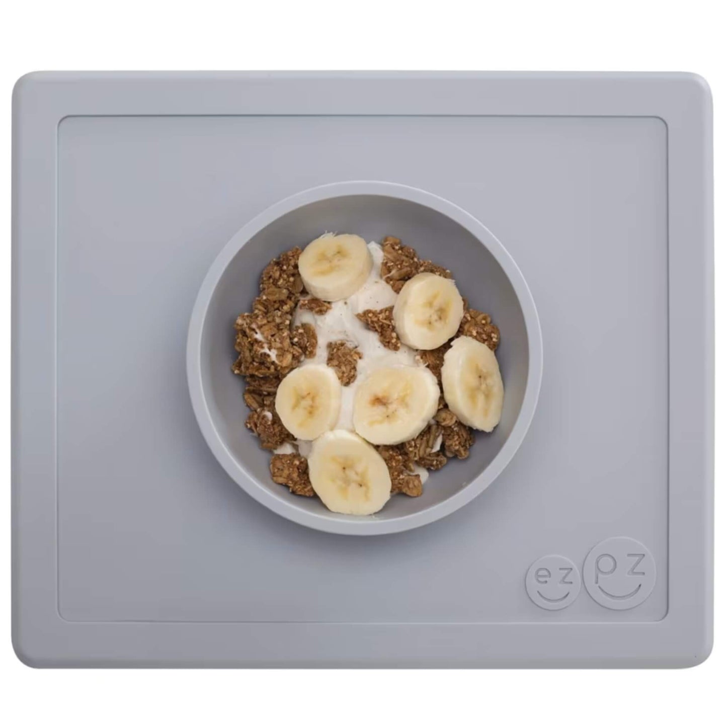 Grey silicone placemat with built-in bowl neatly filled with yoghurt, granola clusters and banana slices arranged on top.