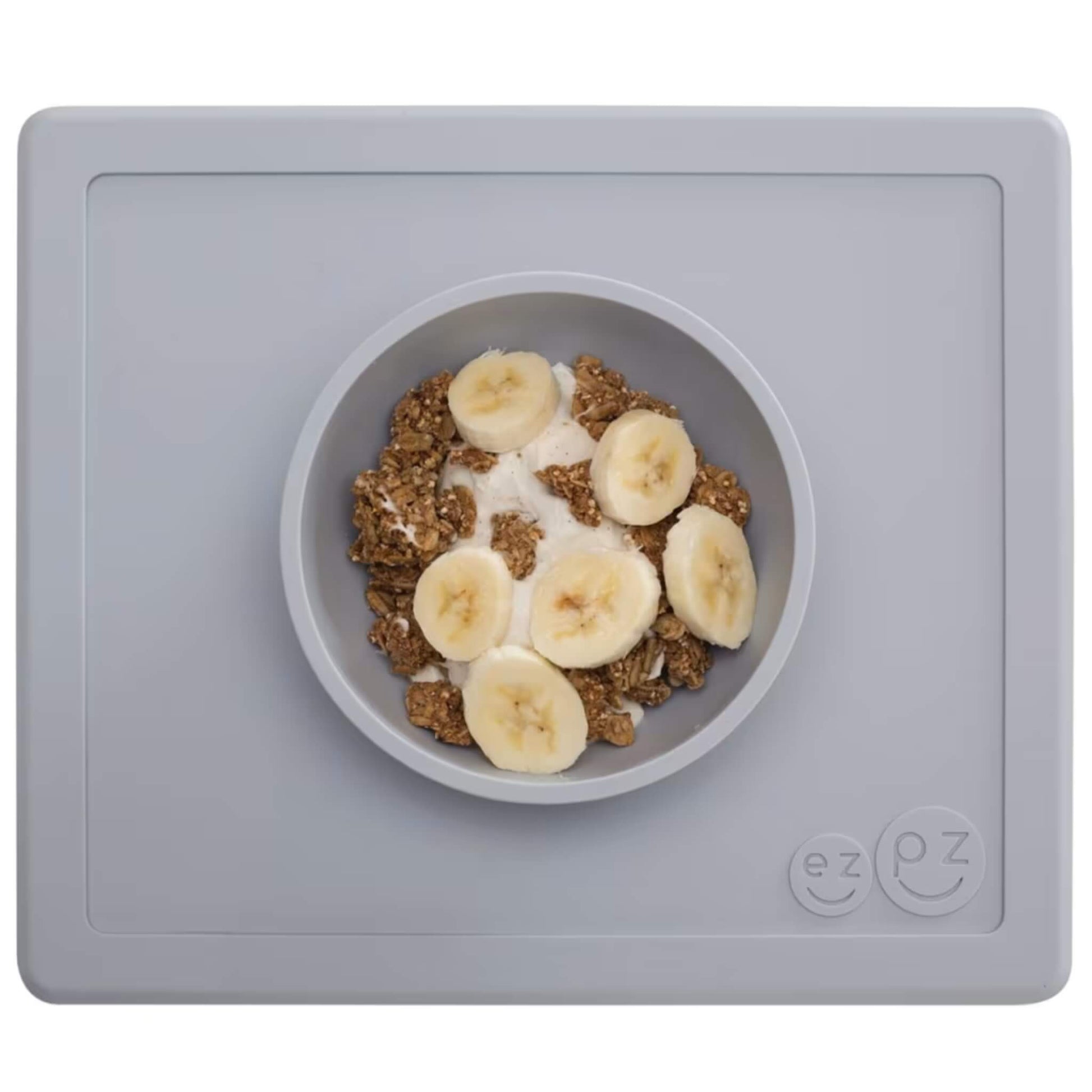 Grey silicone placemat with built-in bowl neatly filled with yoghurt, granola clusters and banana slices arranged on top.
