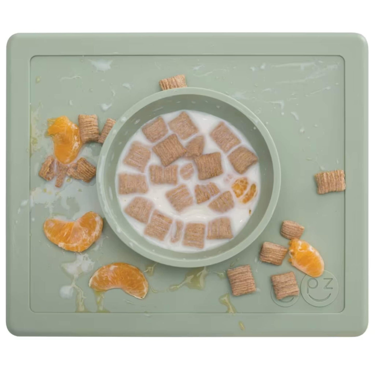 Sage green silicone placemat with built-in bowl filled with milk, cereal squares and orange slices, with food and liquid spilled across the mat surface.