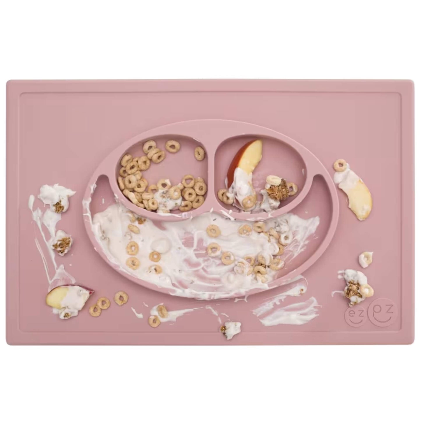 A blush pink silicone placemat and plate filled with cereal, fruit and yoghurt, shown with food spills around the edges after a toddler meal.