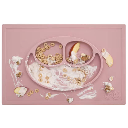 A blush pink silicone placemat and plate filled with cereal, fruit and yoghurt, shown with food spills around the edges after a toddler meal.