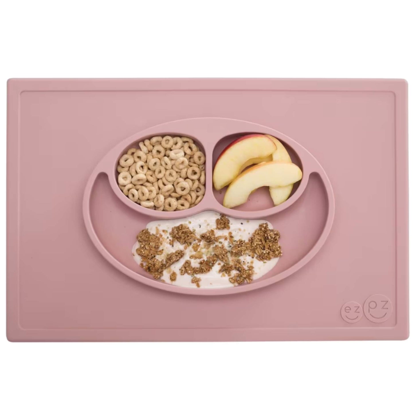 A blush pink silicone placemat and plate set with three compartments neatly filled with apple slices, cereal and yoghurt topped with granola.