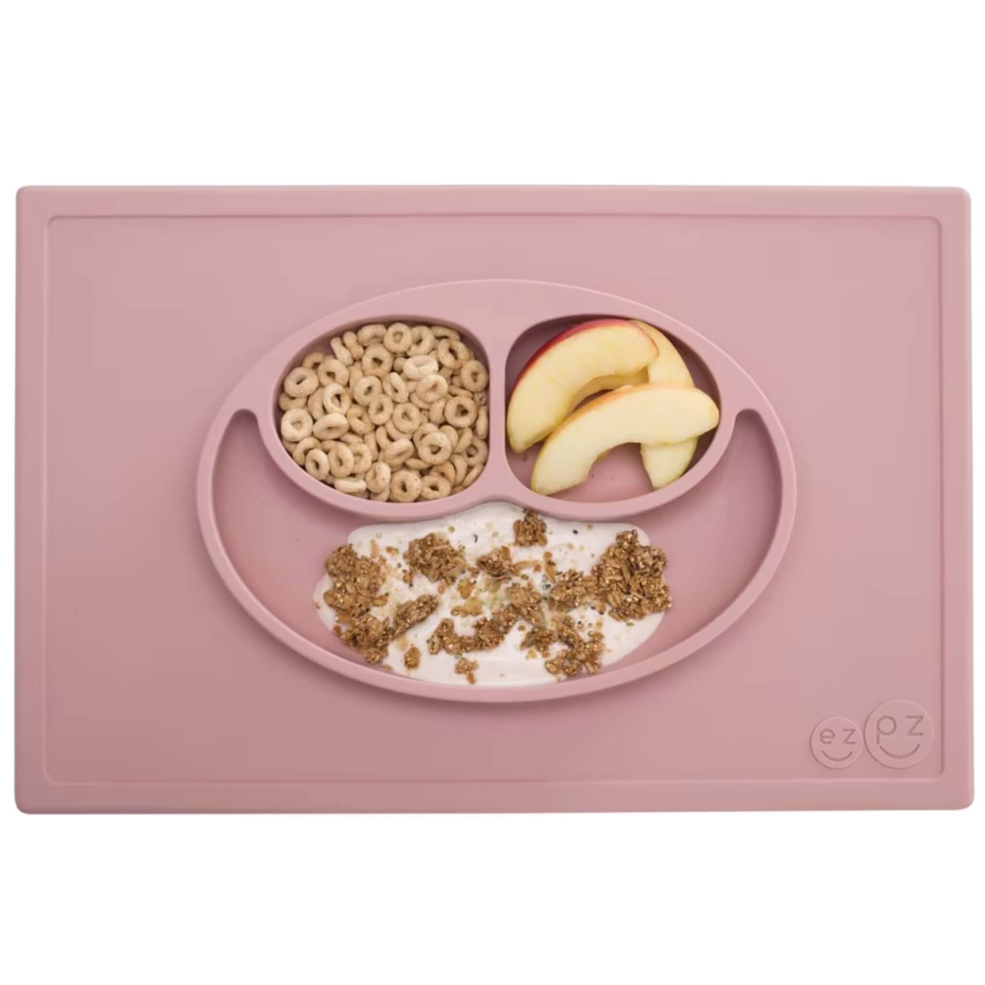A blush pink silicone placemat and plate set with three compartments neatly filled with apple slices, cereal and yoghurt topped with granola.