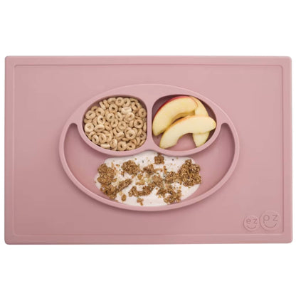 A blush pink silicone placemat and plate set with three compartments neatly filled with apple slices, cereal and yoghurt topped with granola.