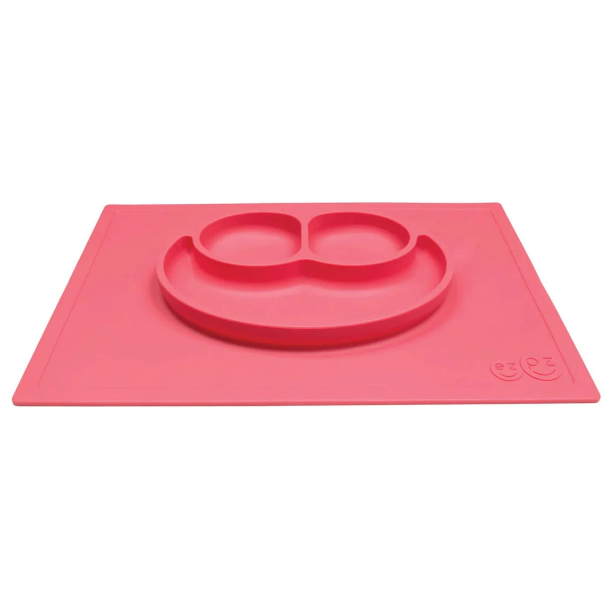 A coral red silicone placemat with an integrated smiley face plate, shown from an angle to highlight its three built-in compartments.