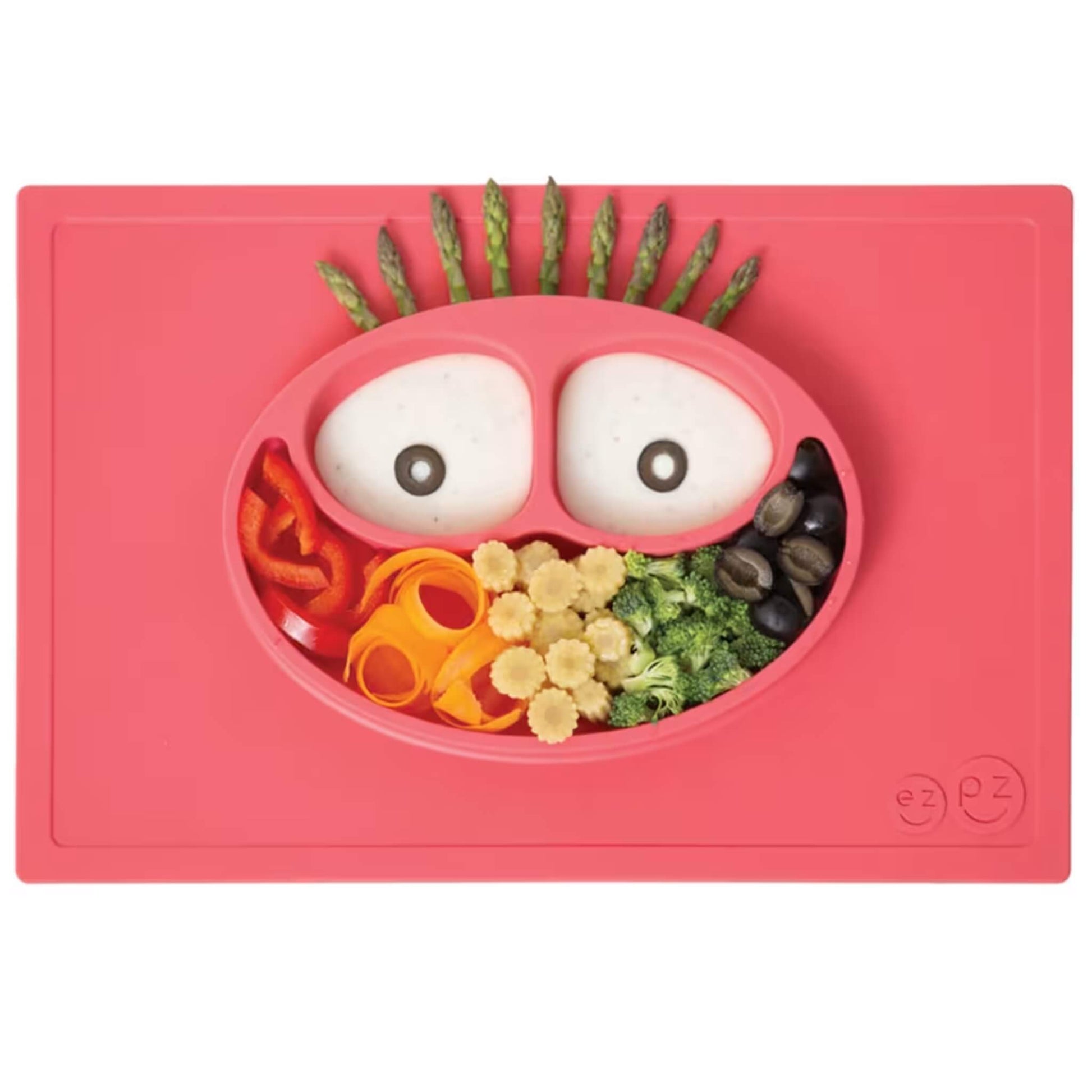 A coral red silicone placemat and plate filled with colourful food arranged in a face design, including broccoli, carrots, peppers and asparagus.