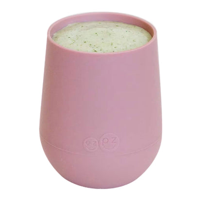 A blush pink silicone training cup shown filled with milk, designed for toddlers learning to drink from an open cup.
